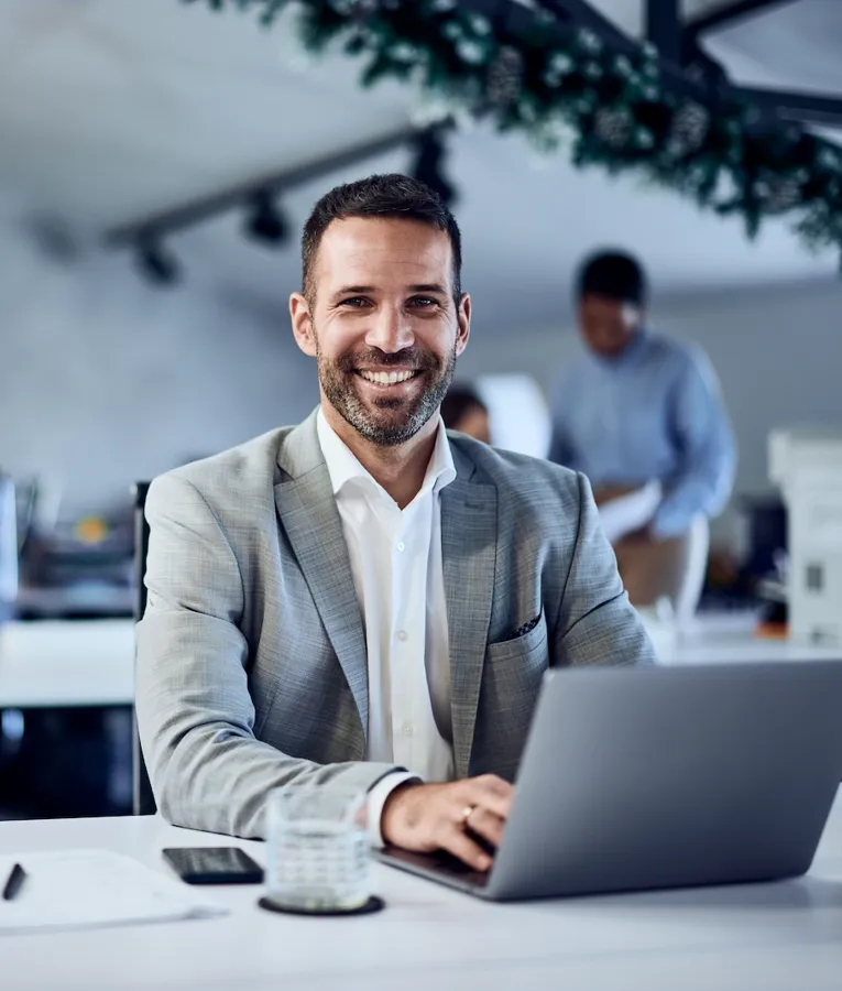 Smilende forretningsmand arbejder på en bærbar computer på et moderne kontor, mens kollegaer samarbejder i baggrunden. Symboliserer arbejdspladsproduktivitet og arbejdsstyrkeoptimering.