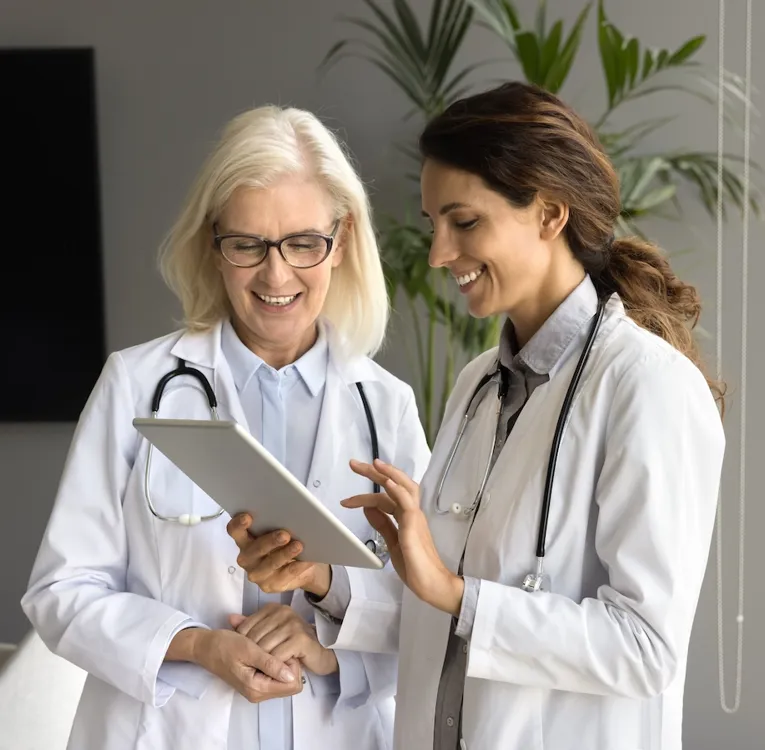 Two female doctors in white lab coats with stethoscopes discuss medical information on a tablet in a modern office with large windows.