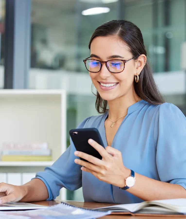 Smilende kontormedarbejder i blå bluse bruger smartphone og bærbar computer ved skrivebordet – symbol på digital kommunikation, hybridarbejde og moderne produktivitet.