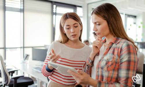Kvindelig ejer af lille virksomhed gennemgår tidsregistrering på tablet sammen med medarbejder på kontor – digital tidsregistrering og vagtplanlægning for små virksomheder med fokus på overblik, løn og medarbejderomkostningsplanlægning.