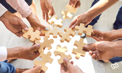 Hænder samles med puslespilsbrikker som symbol på fleksibel vagtplanlægning med blandede ansættelsestyper, hvor fuldtid, deltid og timeløn kombineres effektivt i workforce management og understøttes af præcis tidsregistrering.