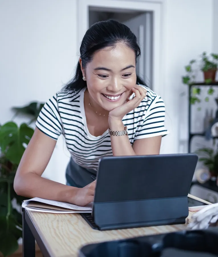 En smilende professionel i en stribet bluse læner sig over et træbord, mens hun arbejder på en tablet med tastatur og deltager i en produktiv samtale på arbejdspladsen.