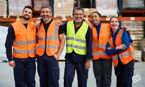 Medarbejdere i logistik- og lagerbranche står samlet på lageret, hvor effektiv vagtplanlægning og workforce management sikrer korrekt bemanding, samarbejde og stabil drift på tværs af skift.