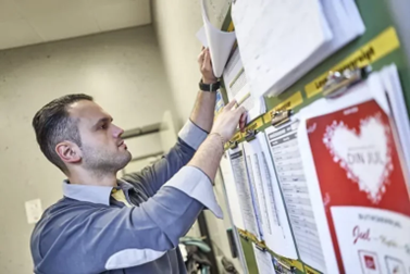 Butikschef gennemgår vagtplan og arbejdsdokumenter på opslagstavle for at sikre korrekt tidsregistrering og bemanding.