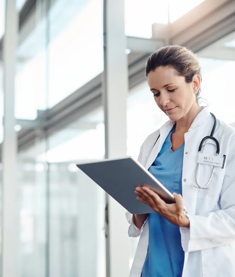 En kvindelig læge i hvid kittel med et stetoskop gennemgår omhyggeligt patientdata på en tablet i en lys, moderne hospitalsgang.