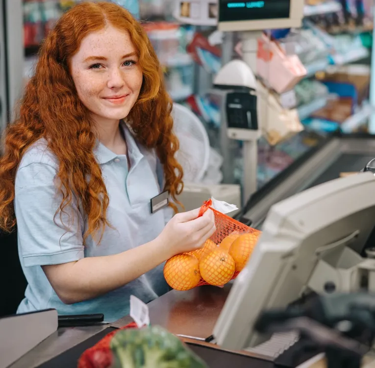 Smilende ung kassemedarbejder i supermarked scanner appelsiner ved kassen – symbol på effektiv bemanding, betjeningssystem og kundeservice.