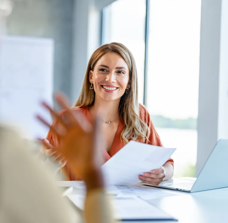 En smilende workforce manager i en rustfarvet bluse holder dokumenter og engagerer sig i en samtale med en medarbejder i et lyst, moderne kontormiljø.