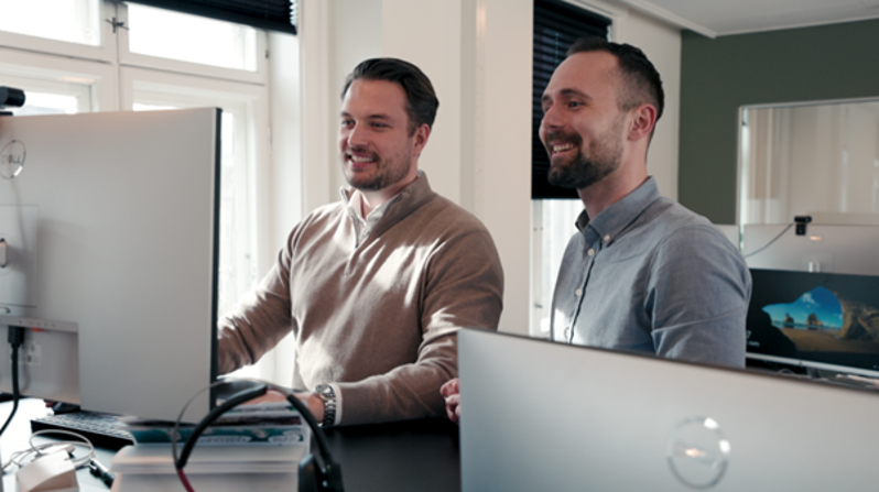 To kollegaer samarbejder foran computerskærm om digital vagtplanlægning og tidsregistrering i et moderne workforce management-system.