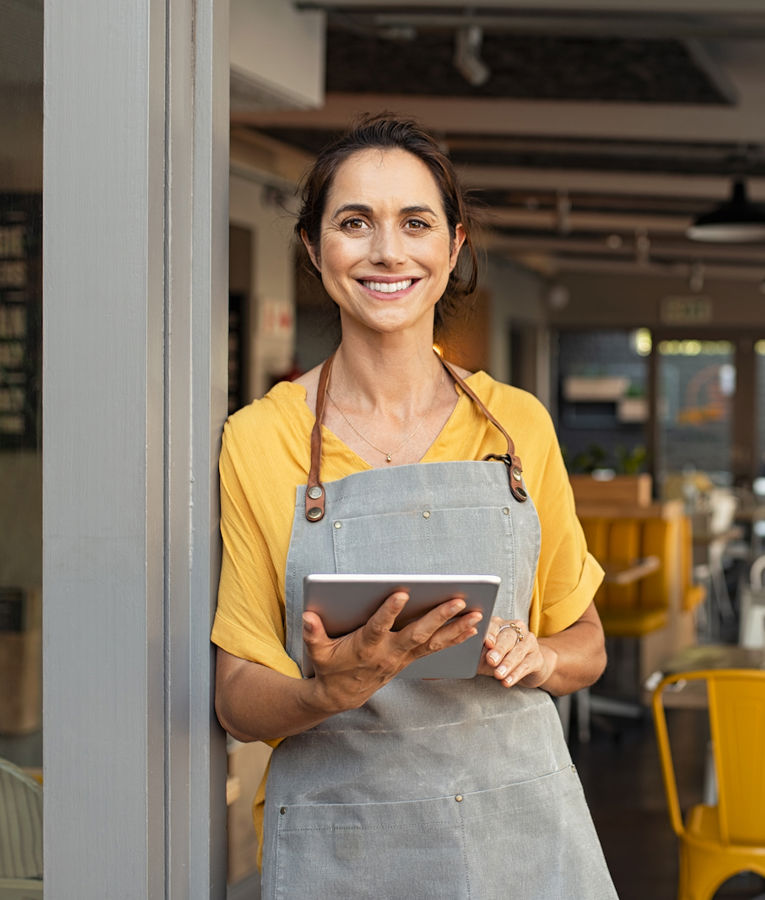 En smilende kvindelig restaurantindehaver eller manager iført en gul bluse og en grå forklæde står ved indgangen til en travl café med en tablet i hånden. Hun ser selvsikker og imødekommende ud, mens hun styrer den daglige drift og kundeservice.