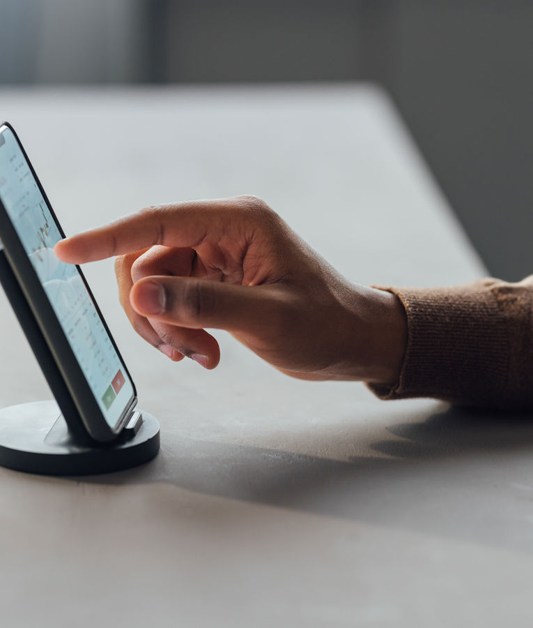 Nærbillede af en person, der bruger en smartphone på en holder og interagerer med en finans- eller dataanalyseapp, der viser diagrammer og grafer. Perfekt til indhold om mobil workforce management, produktivitetsapps eller digitale forretningsværktøjer.