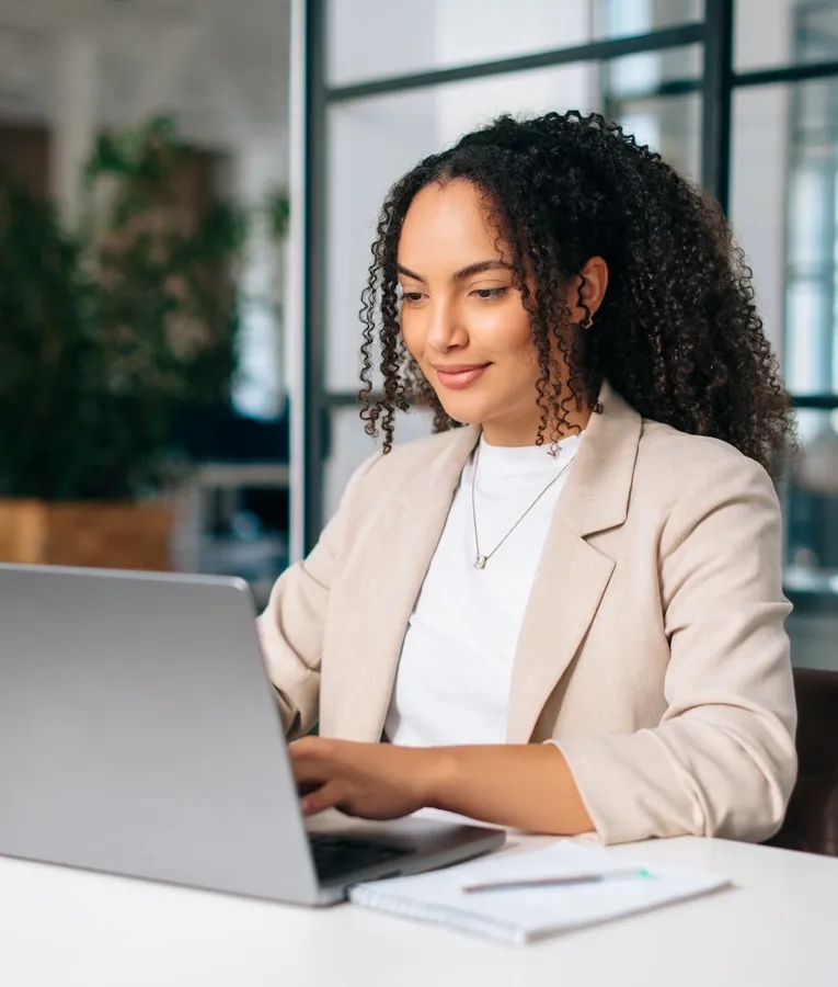 En ung professionel i en beige blazer arbejder på en bærbar computer ved et lyst og moderne skrivebord, omgivet af noter og kontorartikler, hvilket signalerer produktivitet og fokus.