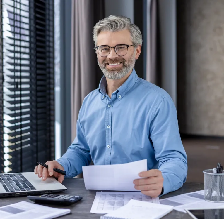 Smilende forretningsmand i blå skjorte arbejder med dokumenter og laptop – symbol på dataanalyse, workforce-rapportering og økonomisk planlægning.