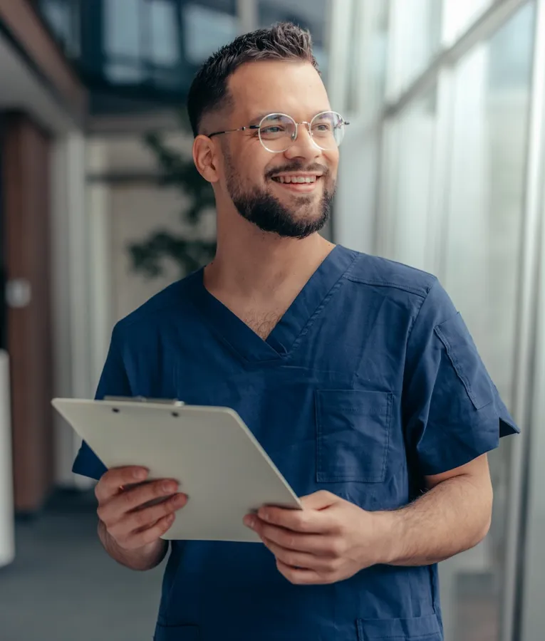En smilende mandlig sygeplejerske i mørkeblå uniform holder en clipboard, mens han står i en lys hospitalsgang.
