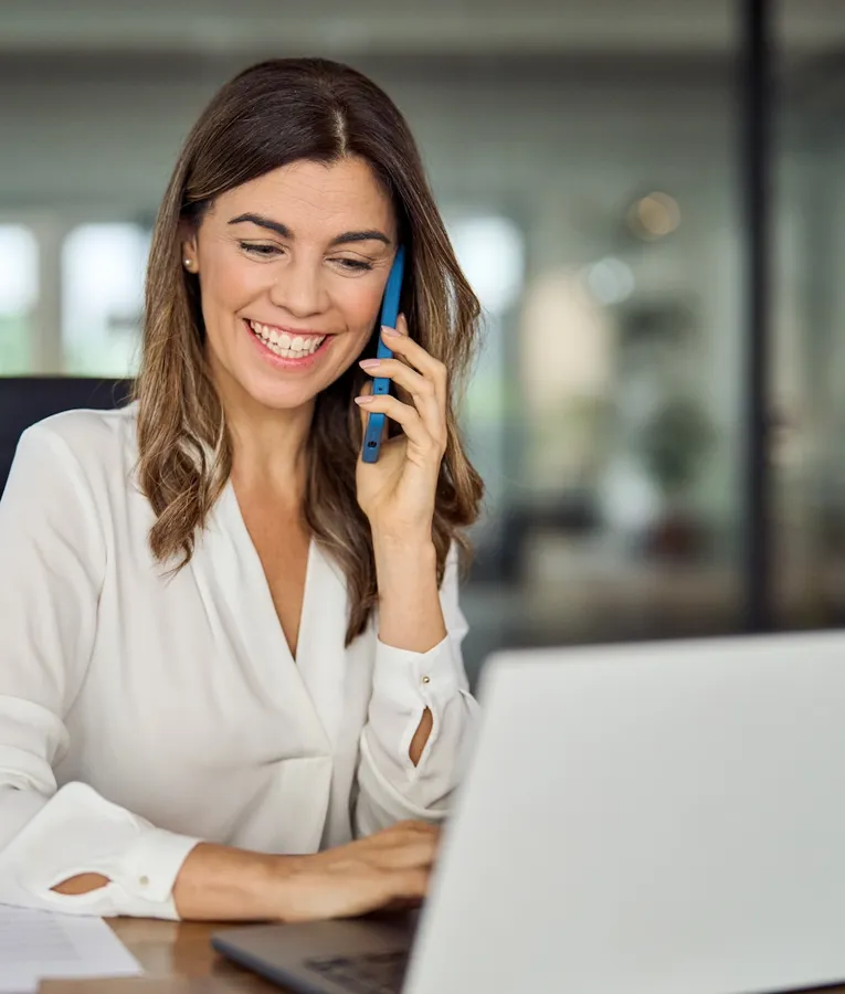 En glad forretningskvinde i en hvid bluse sidder ved sit skrivebord, taler i telefon og arbejder på sin bærbare computer. Billedet symboliserer produktivitet, kommunikation og effektiv arbejdsstyring.