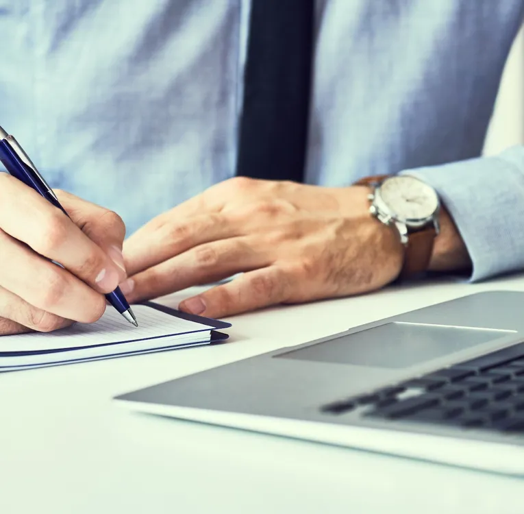 Et nærbillede af en forretningsmand i en blå skjorte og slips, der skriver noter i en notesbog, mens han arbejder på en bærbar computer ved et moderne kontorbord.