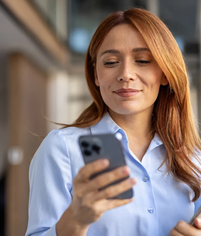 Selvsikker forretningskvinde bruger en smartphone, mens hun holder en tablet, og administrerer opgaver eksternt. Symboliserer mobil arbejdsstyring og digital effektivitet.