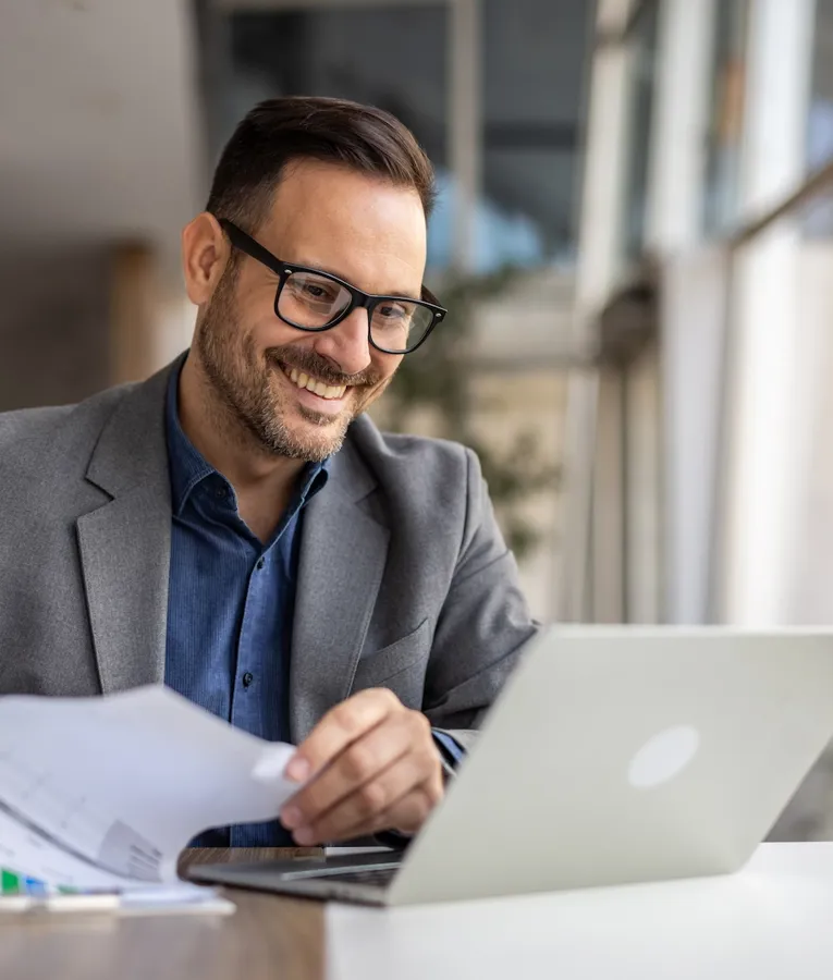 En smilende forretningsmand i en grå blazer og briller gennemgår arbejdsstyrkeanalyse på en bærbar computer, mens han holder finansielle rapporter på et moderne kontor.
