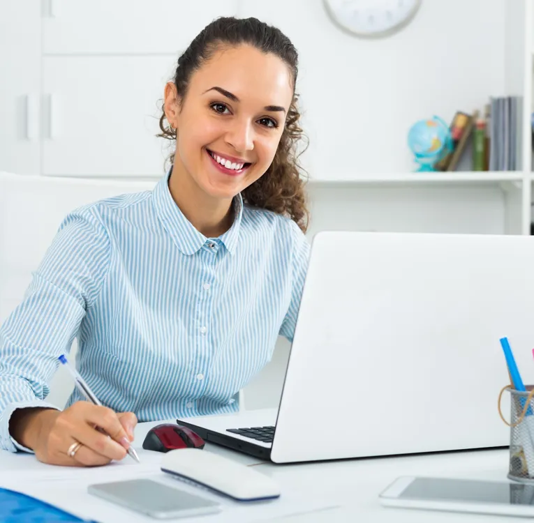 Glad kontormedarbejder i stribet skjorte tager noter ved skrivebordet med laptop – symbol på planlægning, koordinering og digital administration.