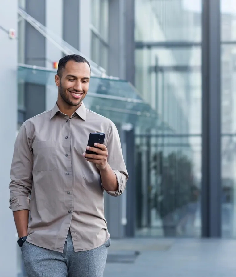 En smilende professionel i en beige skjorte går udenfor en moderne glasbygning og tjekker sin smartphone, hvilket symboliserer en mobil og fleksibel arbejdsstil.